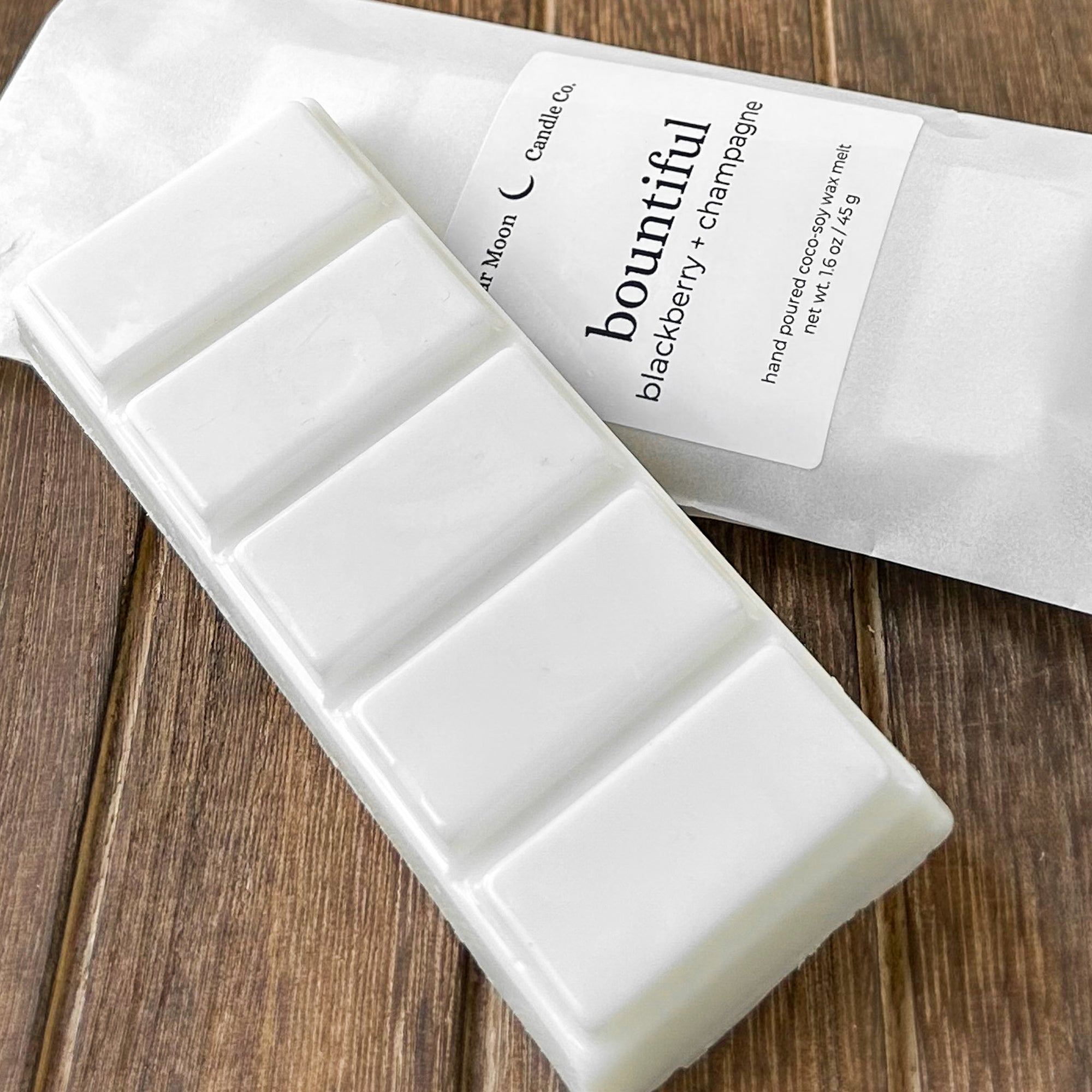 A white soy wax melt labeled 'bountiful' with scents of blackberry and champagne, resting on a wooden surface. The packaging is partially visible behind the wax melt.